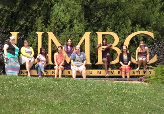 group of women sitting and standing on a large UMBC sign outside